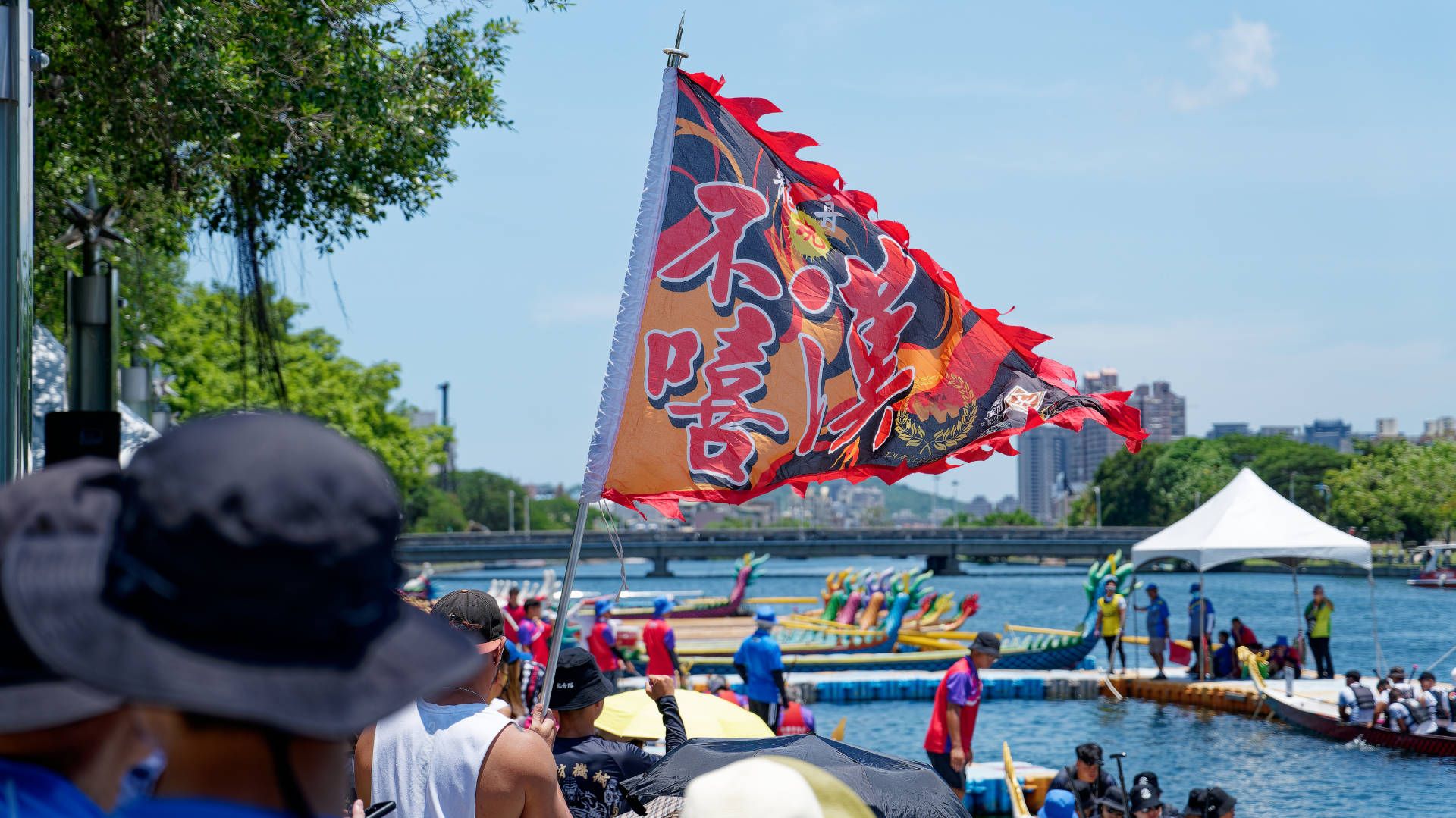 Amongst a crowd of spectators at the dragon boat tug-of-war competition, a man holds a flagpole with a large triangular flag comprising different patterns and Chinese characters.