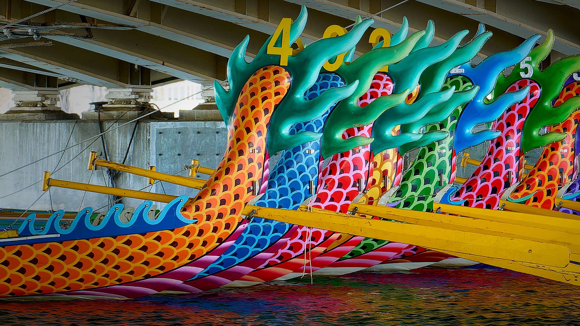 The tails of approximately 10 dragon boats moored under a bridge.