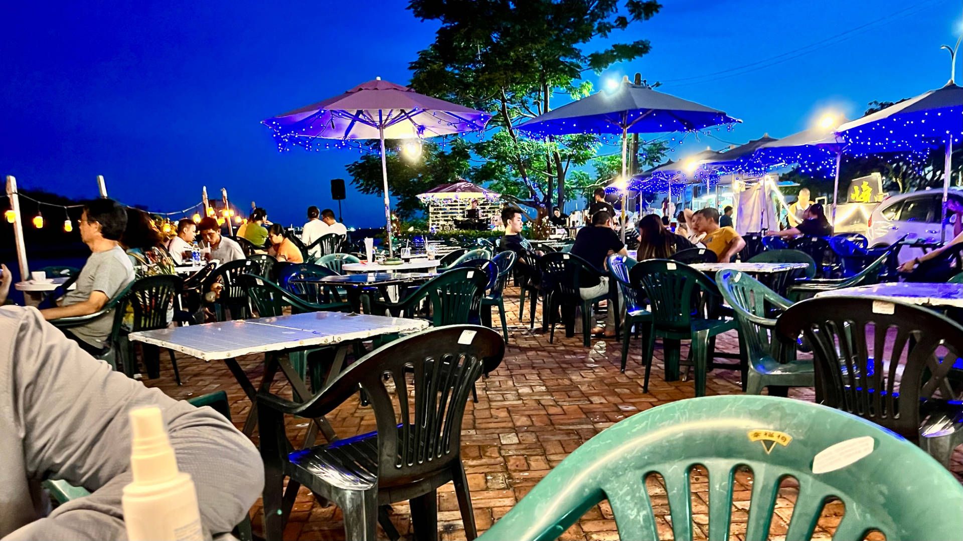 People sitting at outdoor tables, under umbrellas and festoon lights, at a cafe. A musician is singing on a stage in the distance.