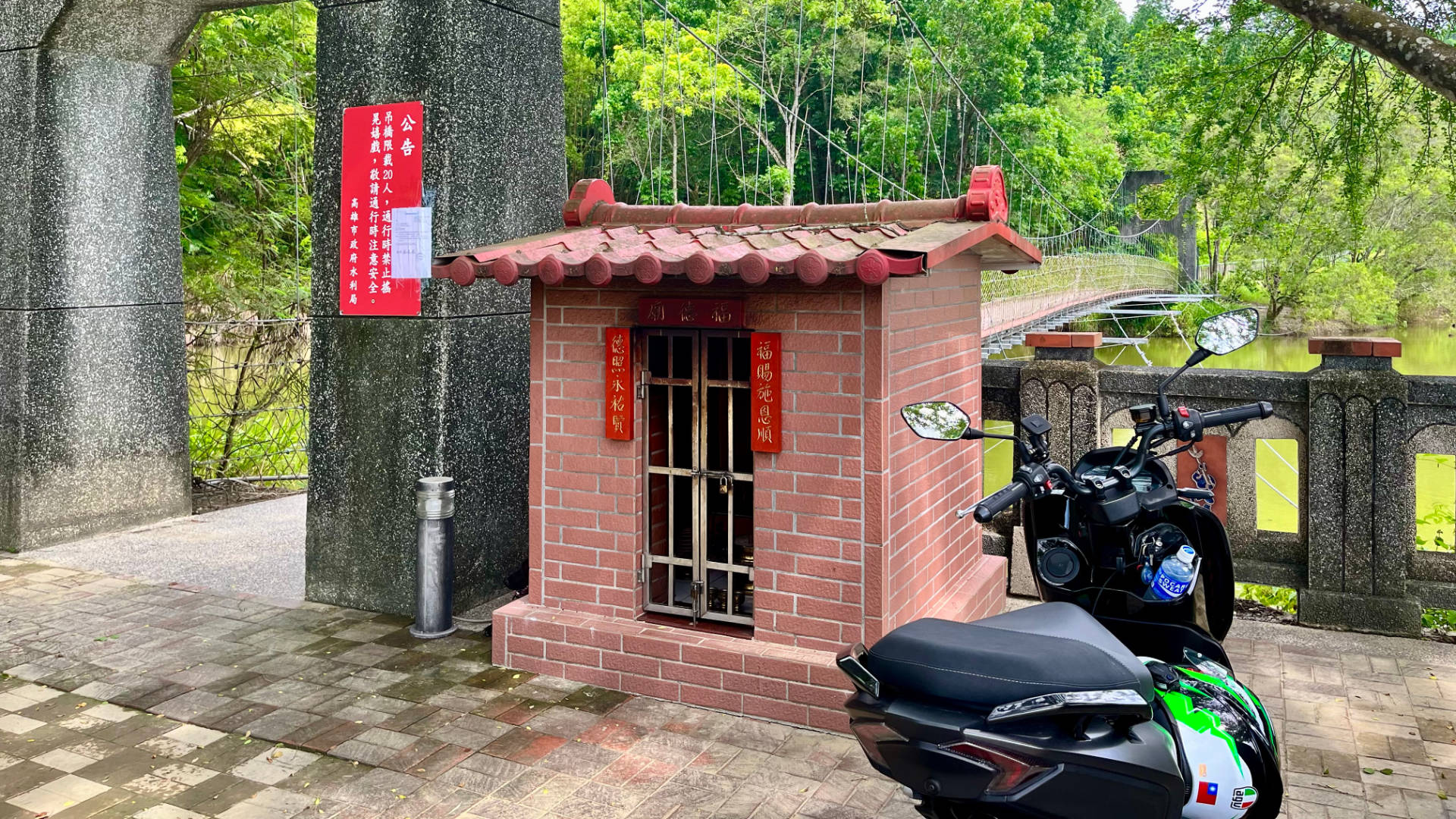 A scooter parked beside a small shrine and a swing-bridge over a green lake, with a forest on the other side.