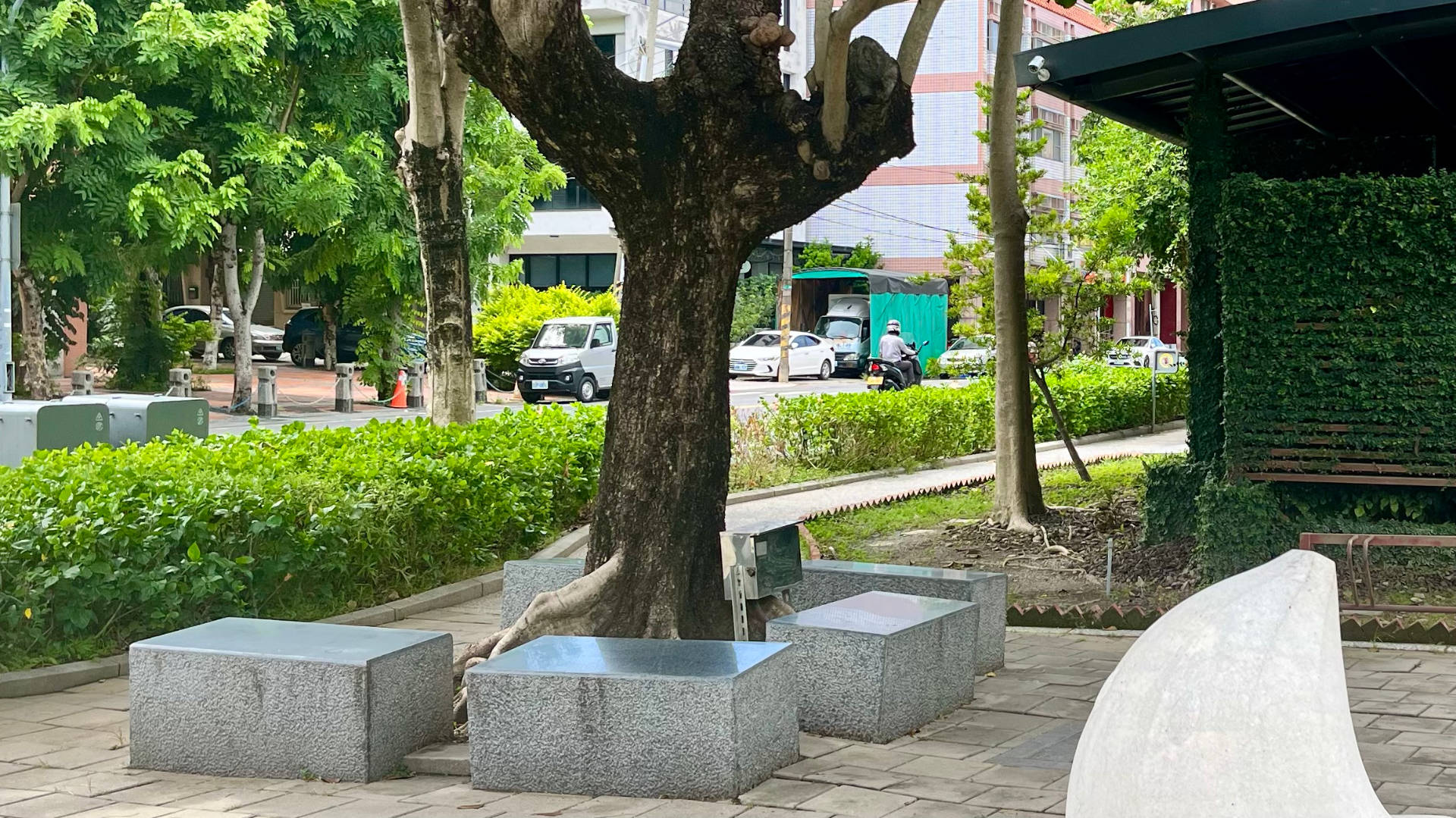 Five short granite benches around a tree, with a public toilet building (and security camera) a few meters away.