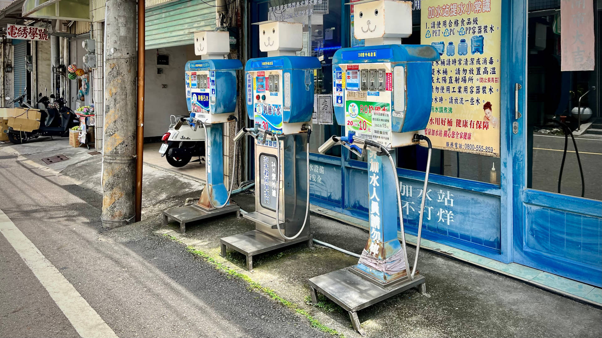 Three drinking water machines on the side of the road. Each looks a little like a petrol bowser, with a rectangular smiling face on the top. The machines are coin-operated.