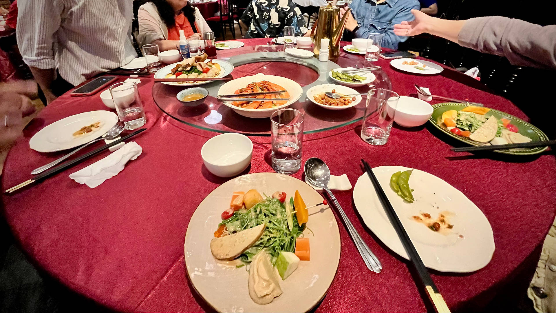 A round banquet table with multiple dishes. There are approximately 10 people seated around the table (their faces are cropped out of frame).