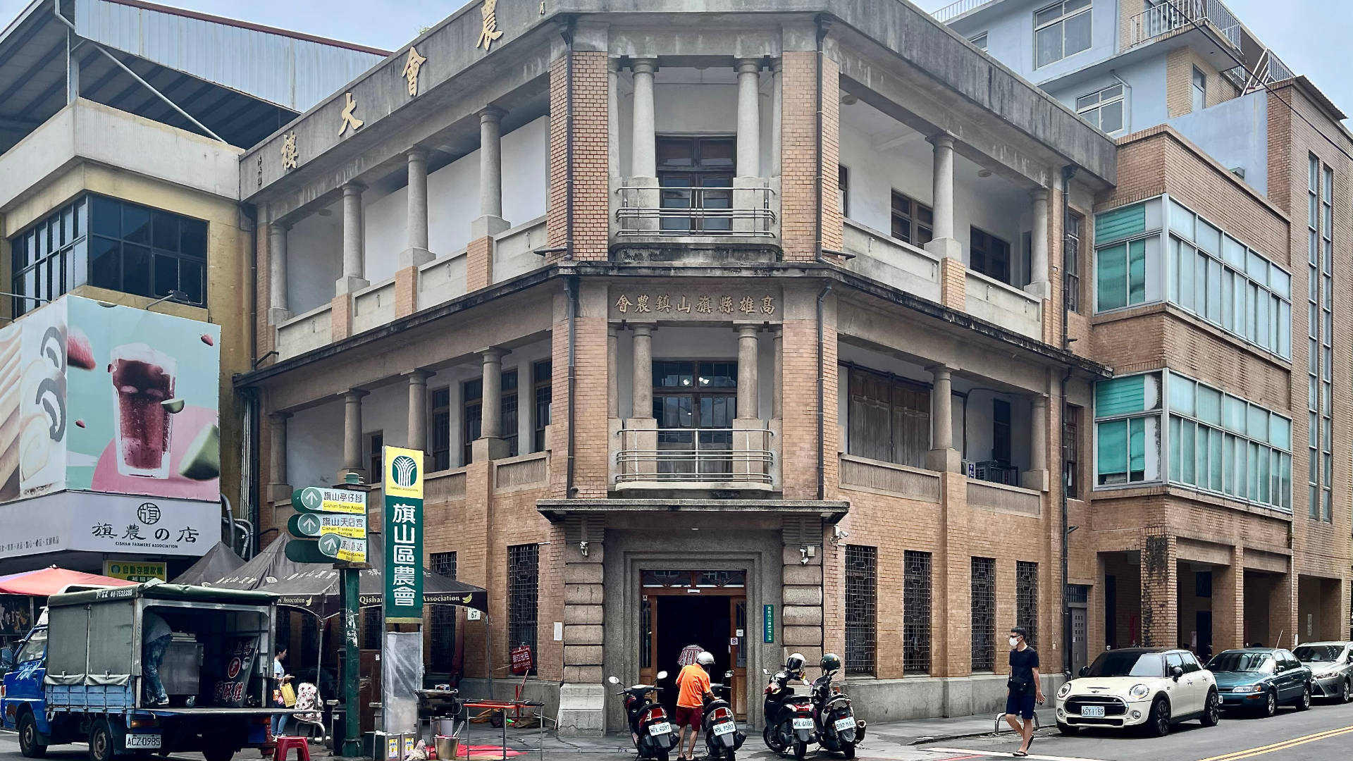 A historic three-story brown brick-and-concrete corner building with four scooters parked outside. Cars are parked on the street to the right, and a delivery van on the street to the left.