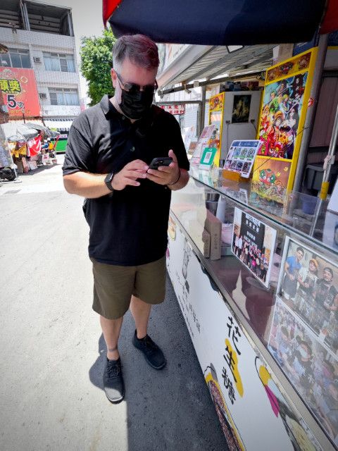 A man standing in the shade outside Moose Peanut ice-cream shop, wearing a mask and sunglasses, and looking at his phone.
