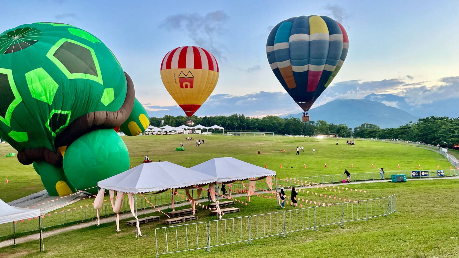 In the foreground, a large turtle-shaped hot air balloon is partially inflated, while two conventionally-shaped balloons are flying behind it. Each is around 10 meters off the ground.