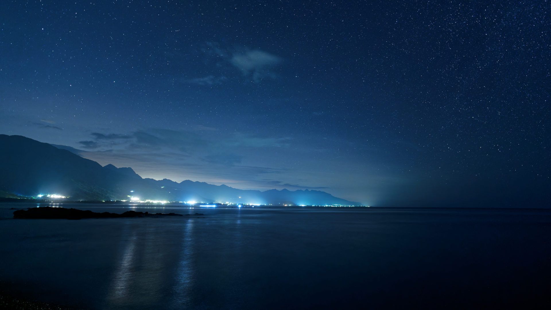 A long-exposure photo of the sea at night, with the lights of coastal towns in the distance. There are stars in the sky. The sea is calm.