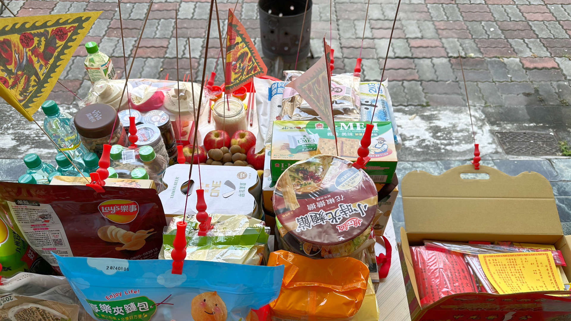 A table of food offerings on a sidewalk in Taiwan. The table is heaped high with food and drinks and burning incense sticks. On the far side of the table, a metal cylinder, slightly larger than a bucket, is sitting on the sidewalk.