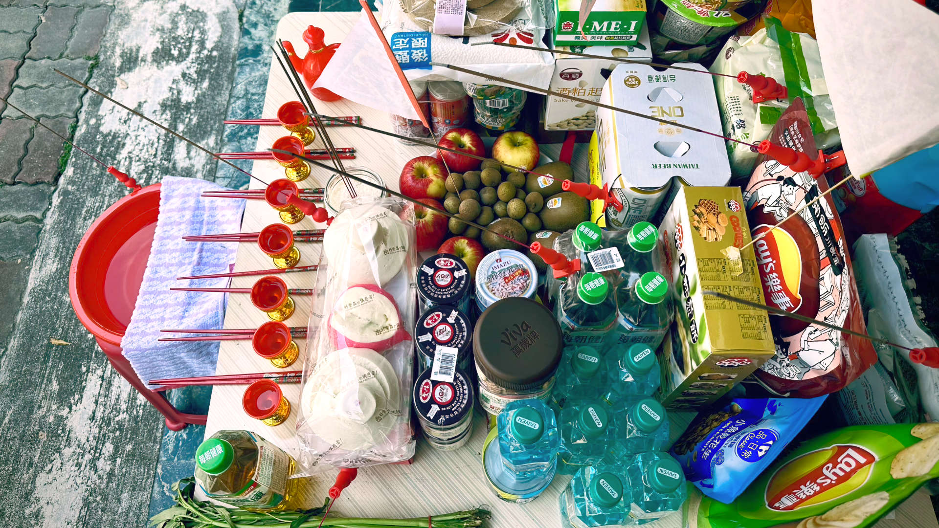 A table, approximately 1 meter square, covered in dry foods, fresh fruit and vegetables, bottled water, beer, and burning incense sticks. A red washbasin is to the left of the table. It is filled with water and has a dry towel resting across the top.