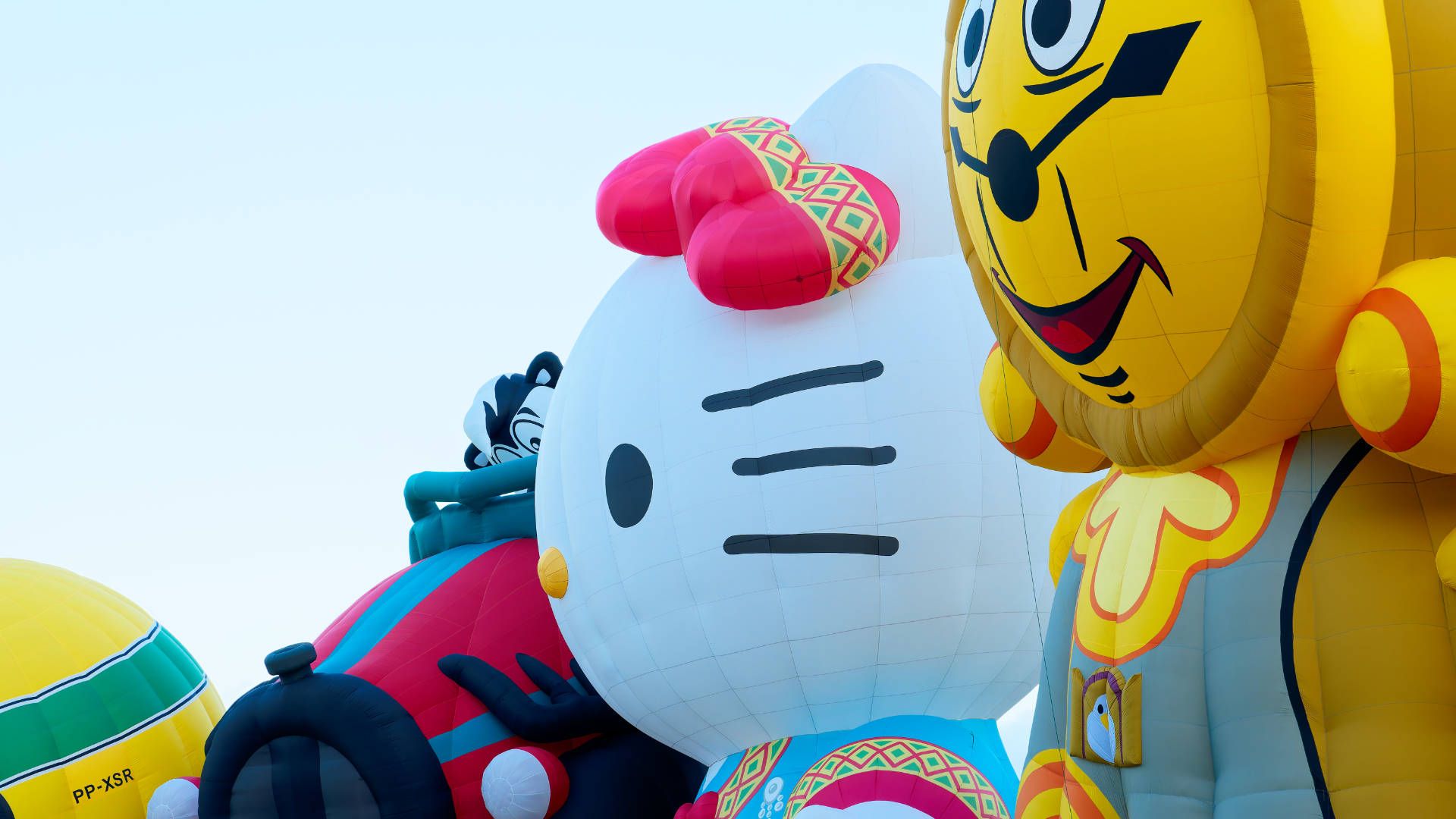Four fully-inflated hot air balloons in a line: A racing helmet, a cartoon skunk in a car, Hello Kitty, and a lion-shaped cuckoo clock.