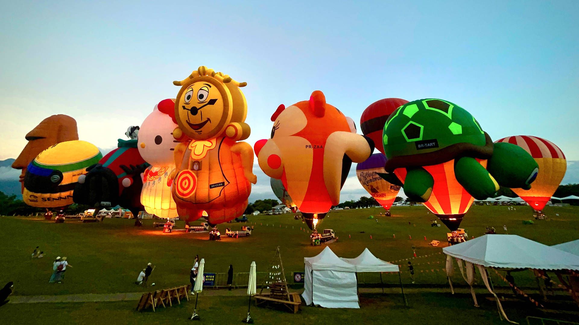 A line-up of 10 hot air balloons, all glowing with their burners ignited. All but one are on the ground.