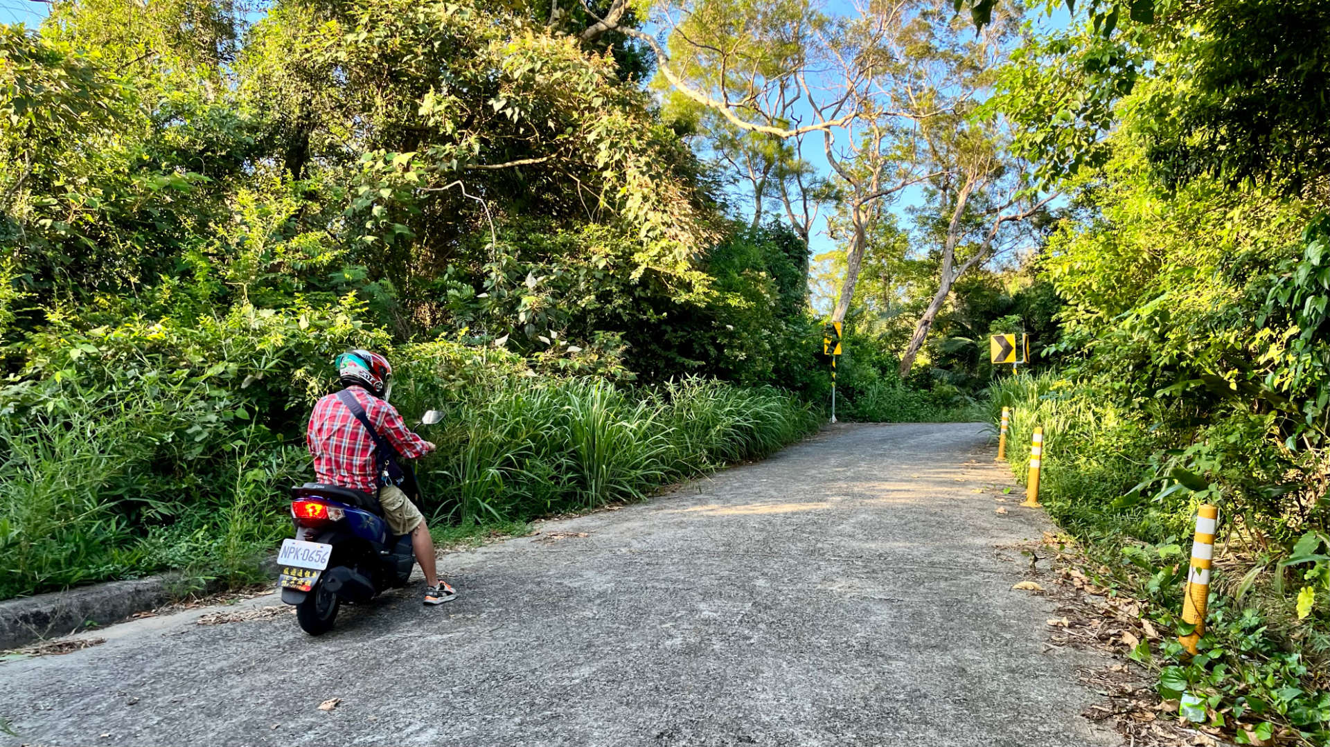 A narrow concrete road leading up a mountain, with dense vegetation either side. One person is stopped on their scooter to the left of the road, looking uphill.