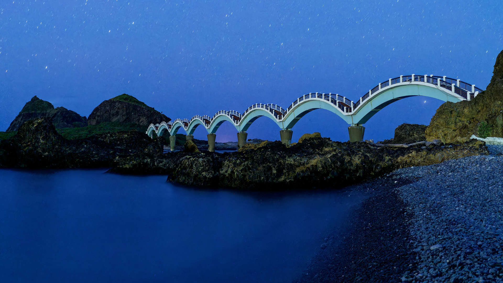 A pedestrian bridge to a small island. The bridge comprises eight arches, each of which people must walk up and over. This is a long-exposure nighttime photograph, with a stony beach in the foreground.