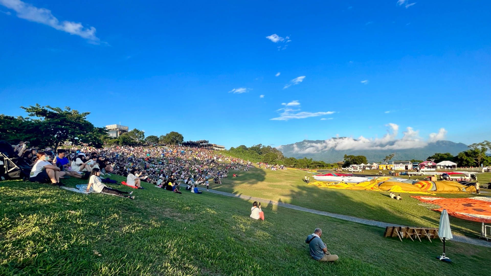 A wide grassy field with a large embankment at one end. A crowd of hundreds of people are sitting on this embankment looking out over a dozen hot air balloons, deflated, and laid out across the grass.