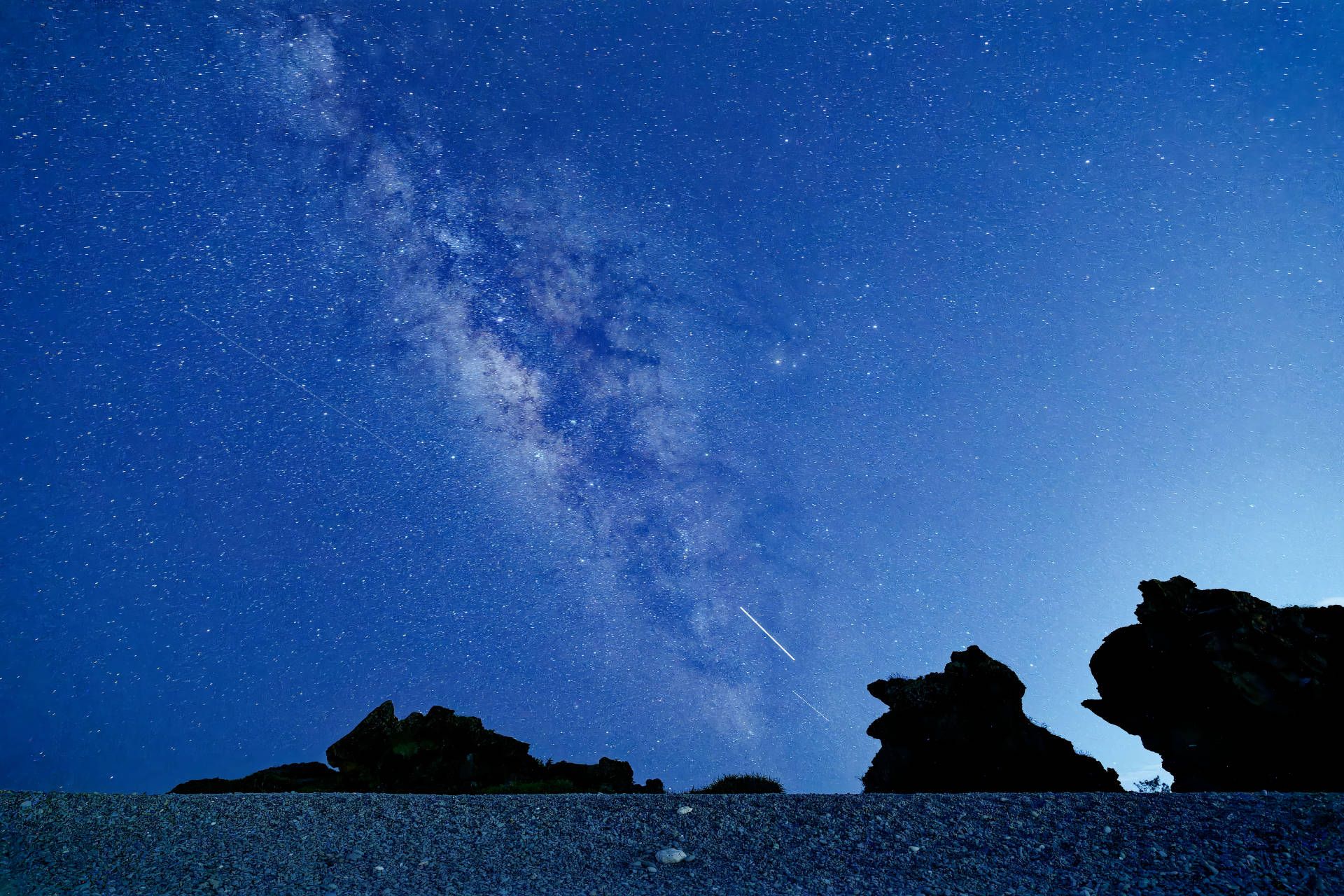 Photo of the milky way extending up from the horizon. In the foreground are the silhouettes of some large rocks, and then the stony beach.