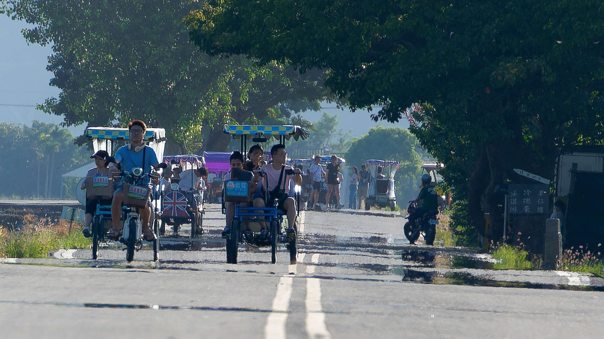 A telephoto shot of perhaps a dozen pedal- and electric-powered bikes and carts on a long country road.