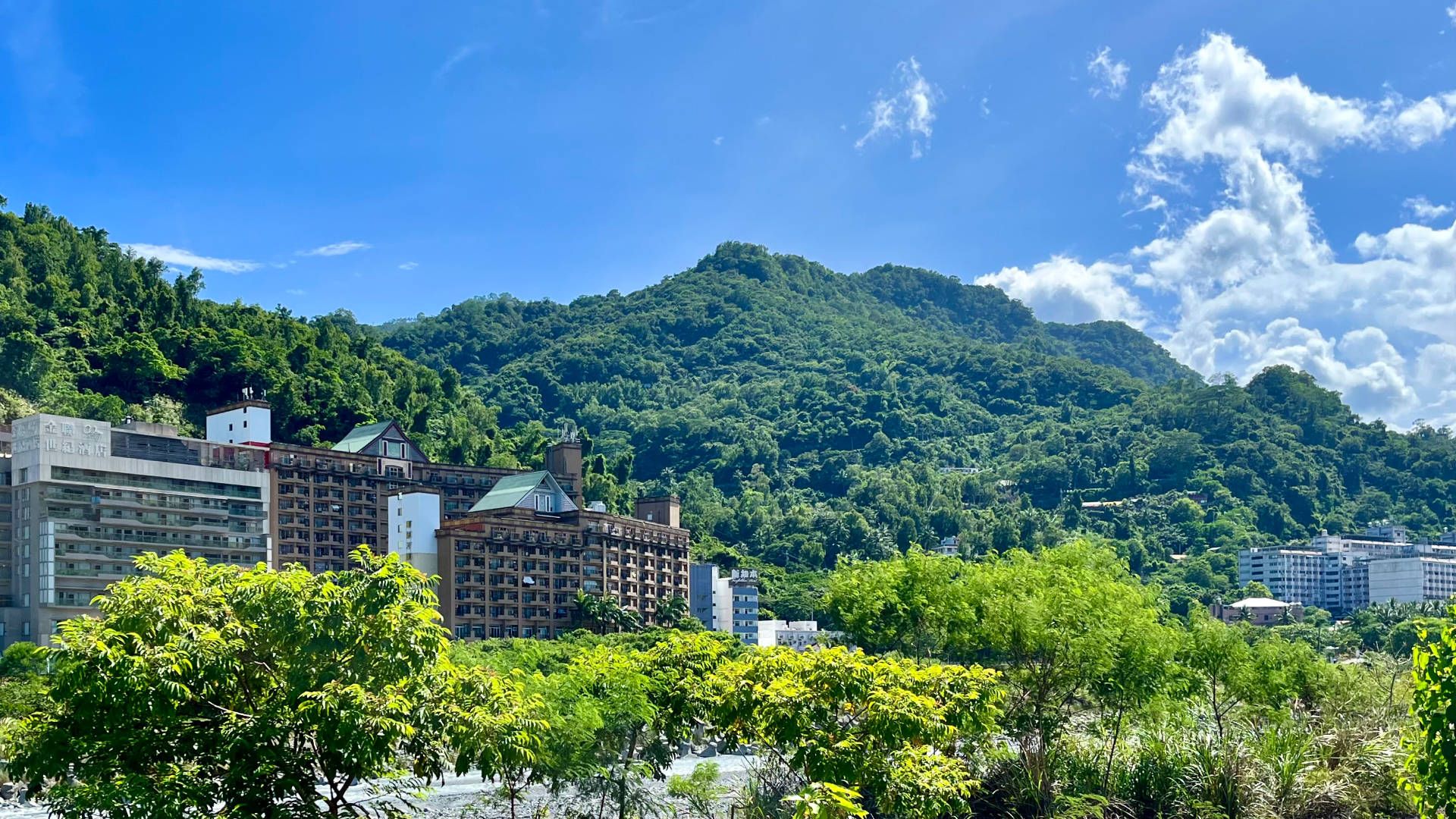 A wide-angle photo of a series of multi-story hotel buildings along a riverfront, with tree-covered mountains in the background.