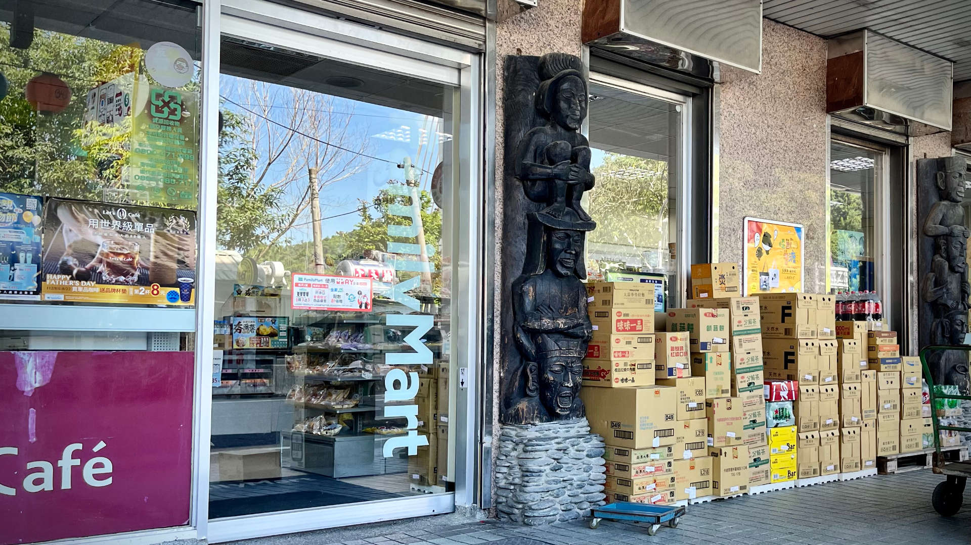 The entrance of a Family Mart convenience store. To the right of the entrance, a wooden sculpture of human-like figures is stacked vertically on a stone base. Next to this, are dozens of boxes of drinks being stored on the pavement.