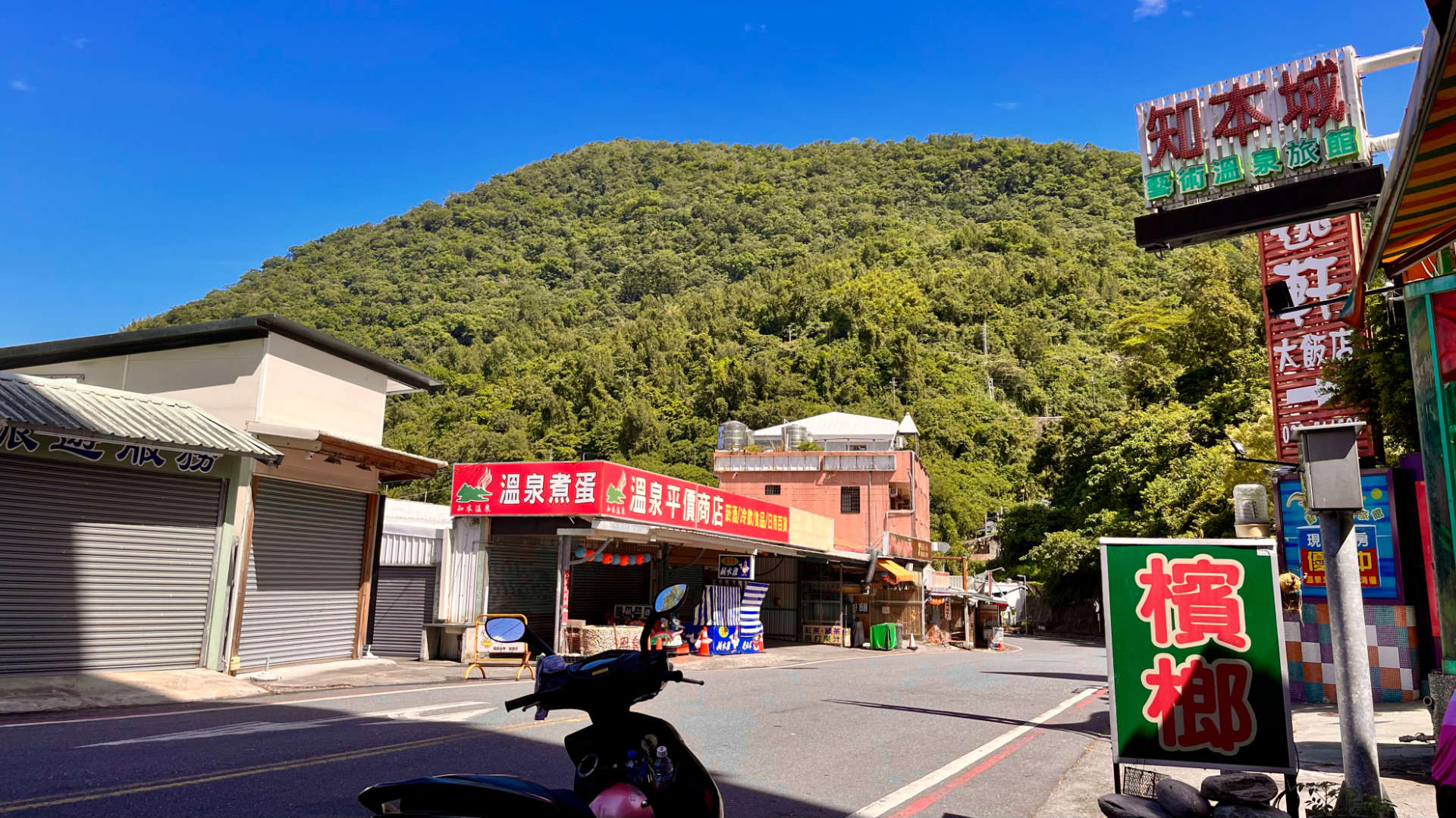 A scooter in the foreground and a quiet streetscape behind, with mountains beyond that. There are no people, and three or four visible shops that look empty.