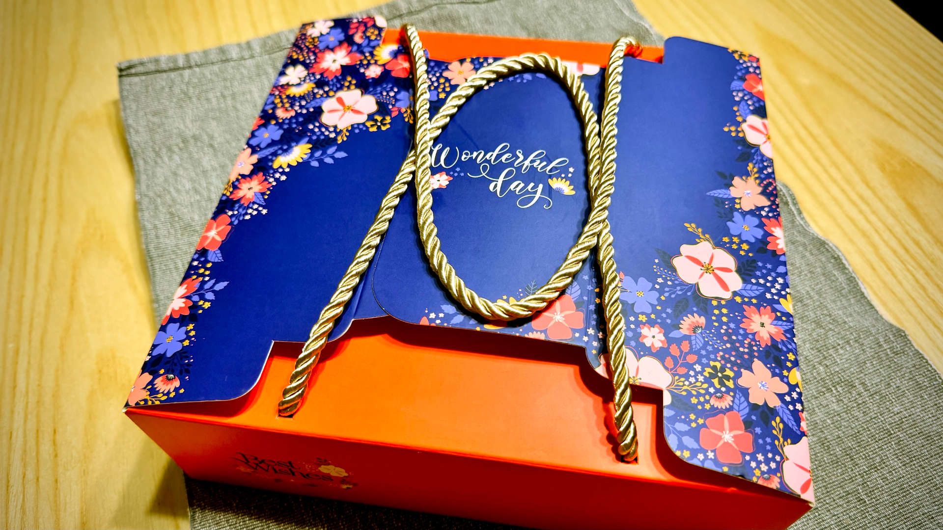 A colorful cardboard box with golden-cord handles. The box is printed with the words ‘wonderful day’ and ‘best wishes’ in cursive script.