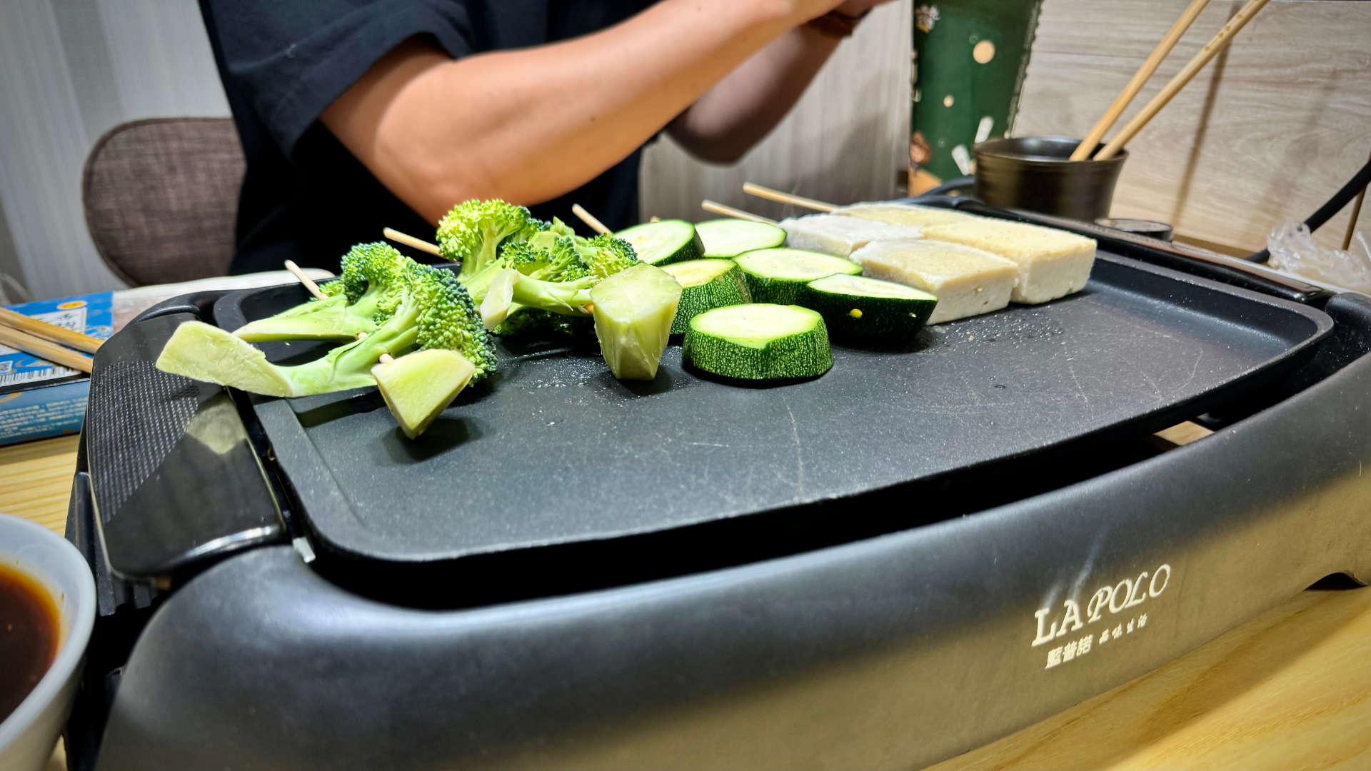 Close-up of an indoor electric grill with skewered broccoli, zucchini, and tofu.