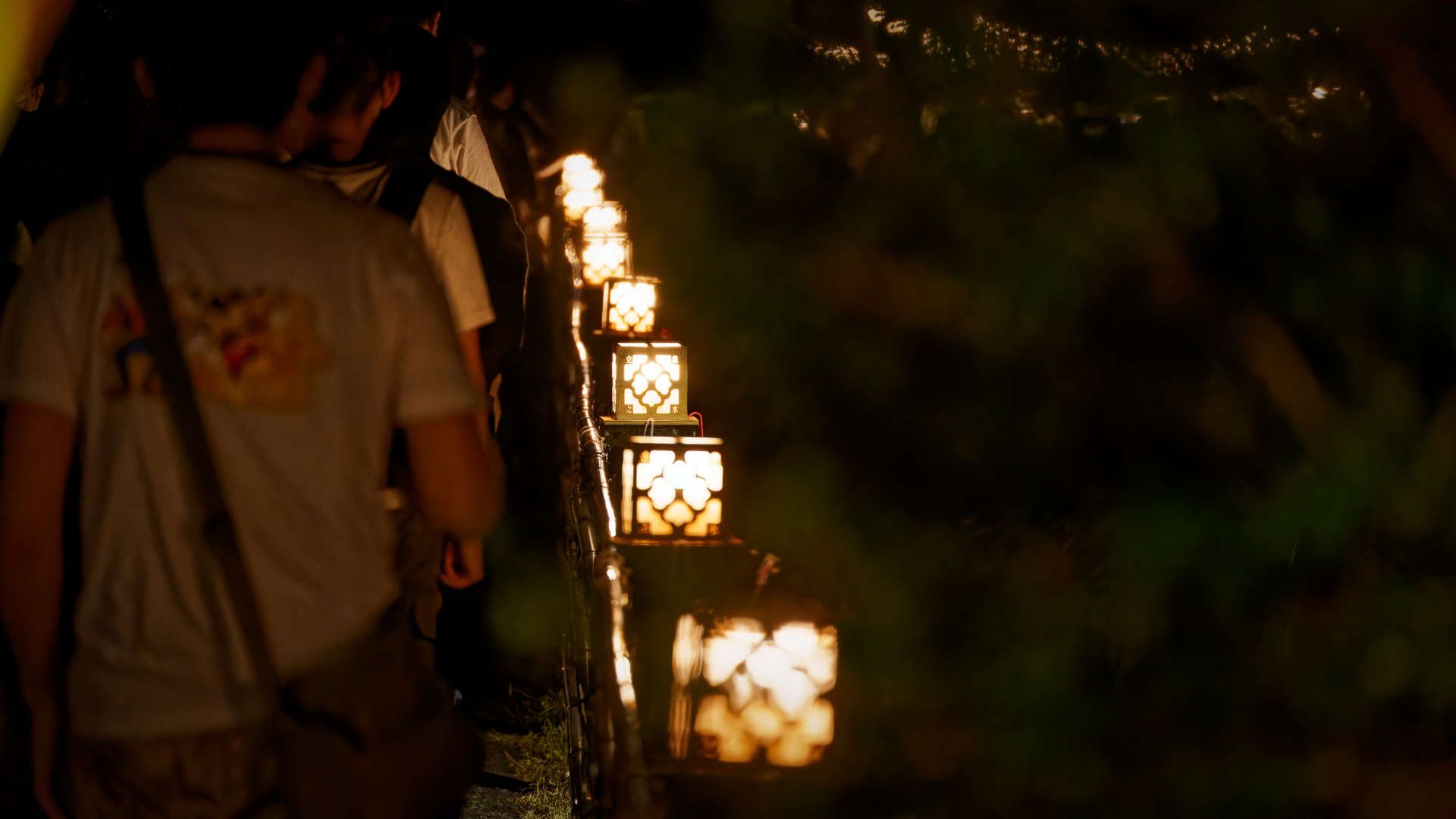 People walking, single file in each direction, across a long footbridge illuminated by small lanterns.