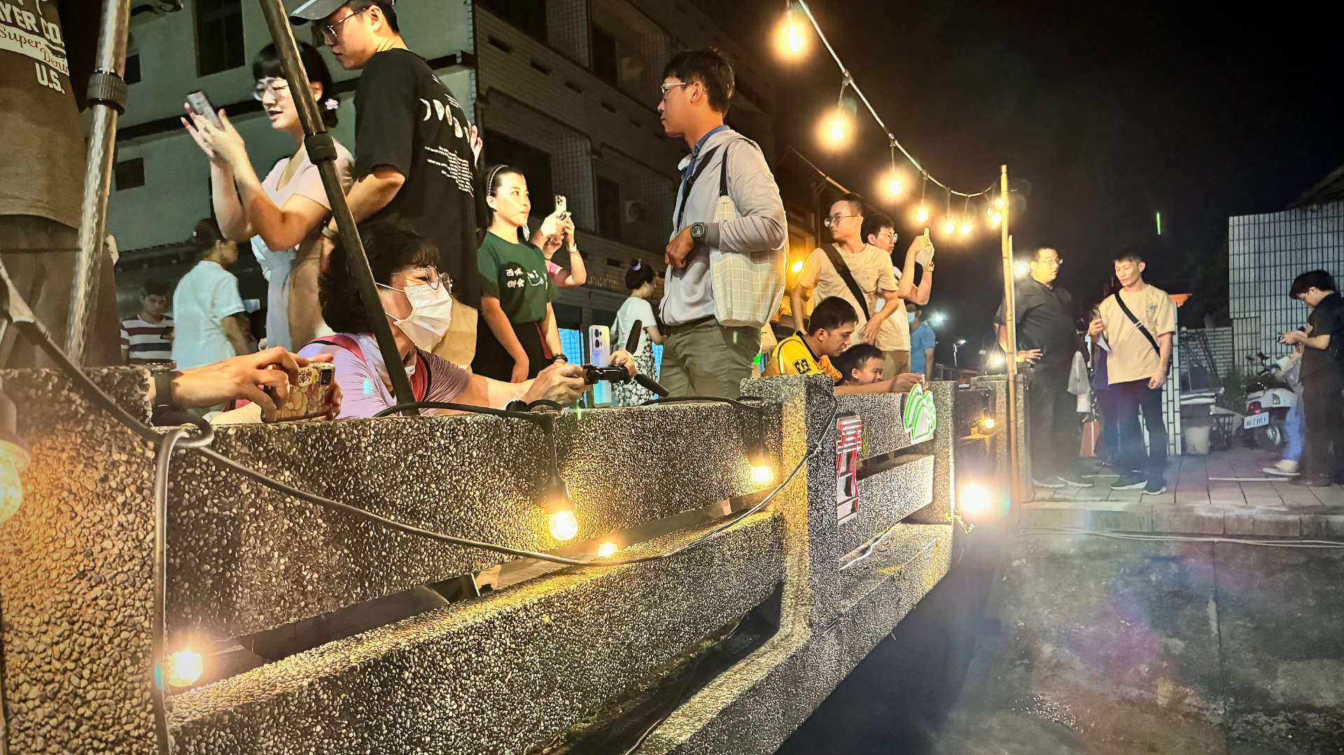 A crowd of people taking nighttime photos on a small concrete bridge, lit by festoon lights.