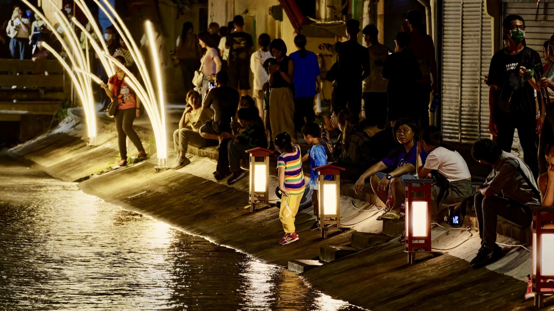 A crowd of people sitting and playing amongst lanterns at night, next to a small urban canal.
