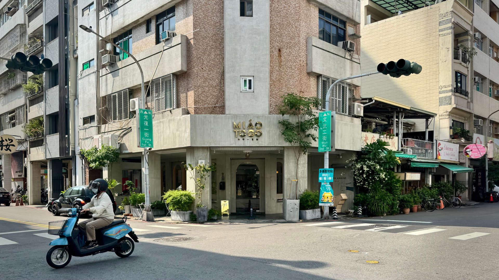 Exterior view of the cornerside ‘Miao To Go’ cat cafe. A woman is driving a scooter past the cafe, which is on the ground floor of a multi-story building on a quiet street corner.