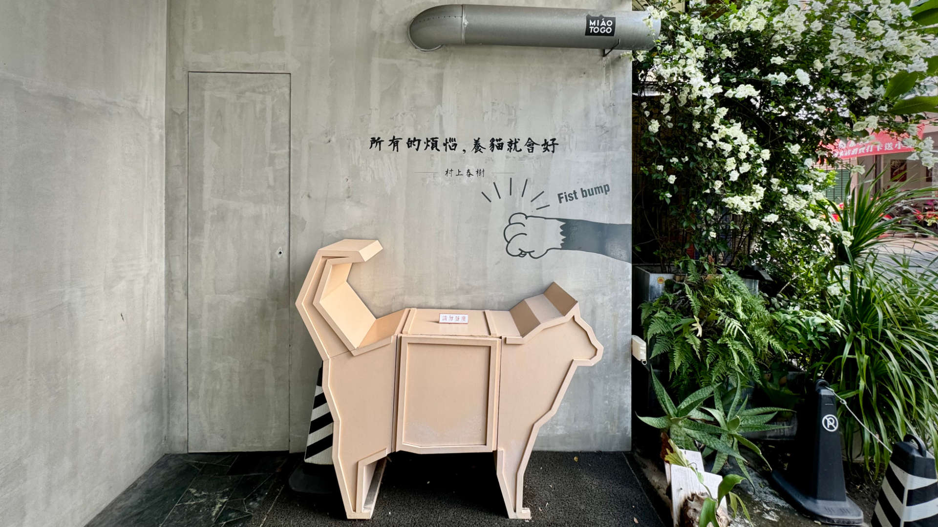 A waist-height wooden cat sculpture, below a concrete wall with Chinese writing on it. Beside the Chinese writing is a cat paw and the English phrase “Fist bump”.