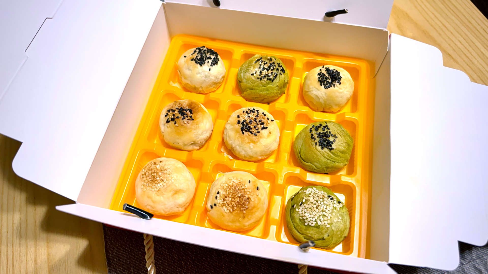 An open cardboard box. Inside is a plastic tray covered in nine round mooncakes. The mooncakes are either green or pastry-colored, and topped with small seeds.