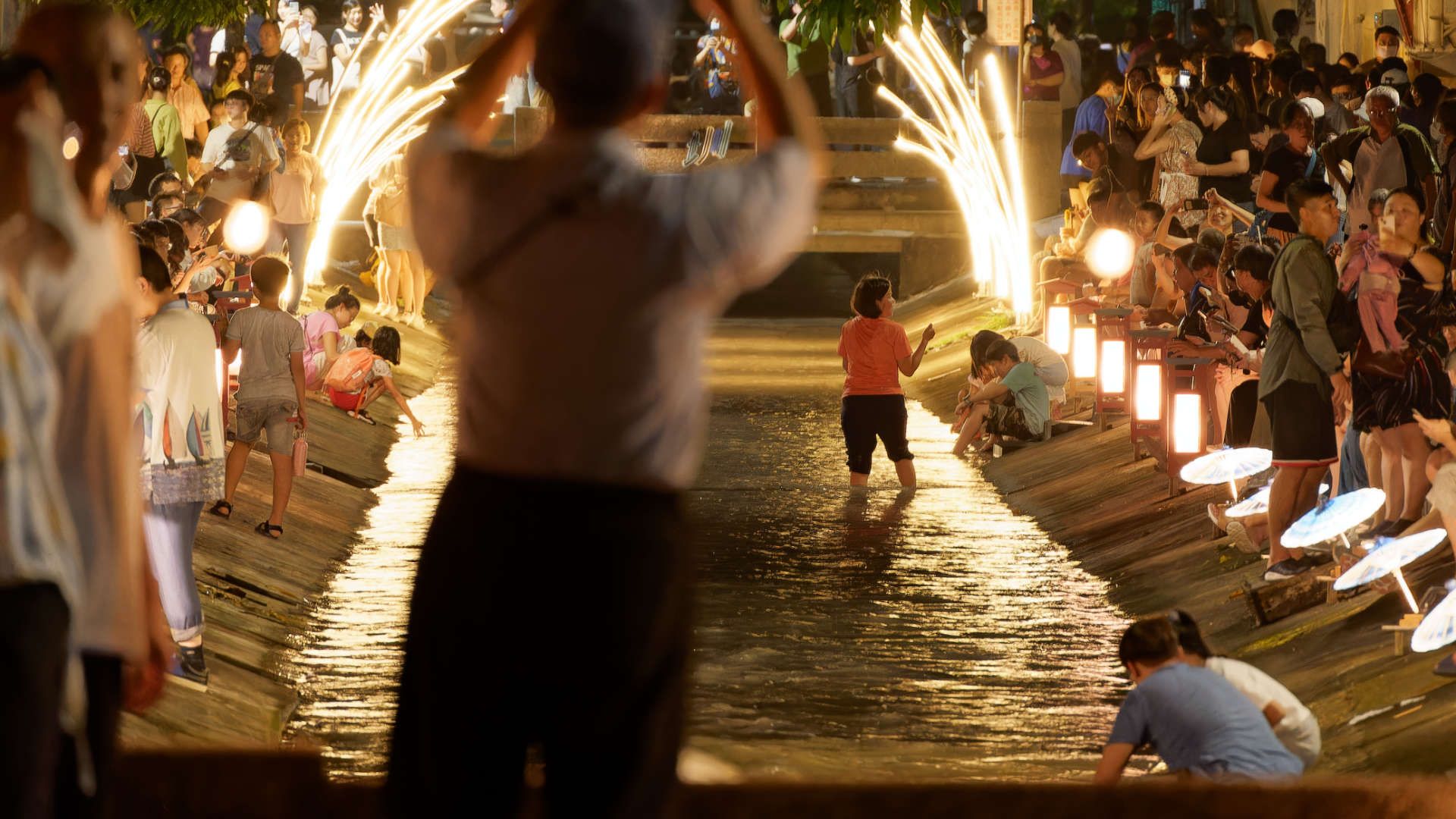 Crowds of people alongside a Meinong canal at night, lit by canal-side lanterns. A few people are in, or touching, the water.