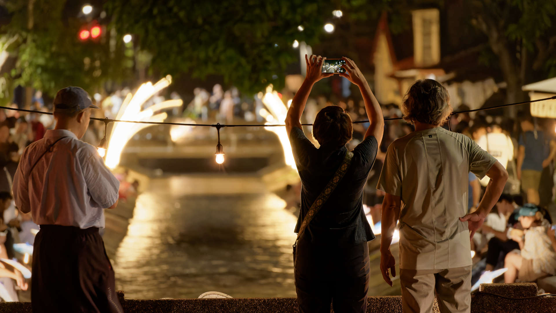 Three people standing on a bridge at night, looking at the view of a lantern-lit canal. One of the people is holding a phone above her head, taking a photo.