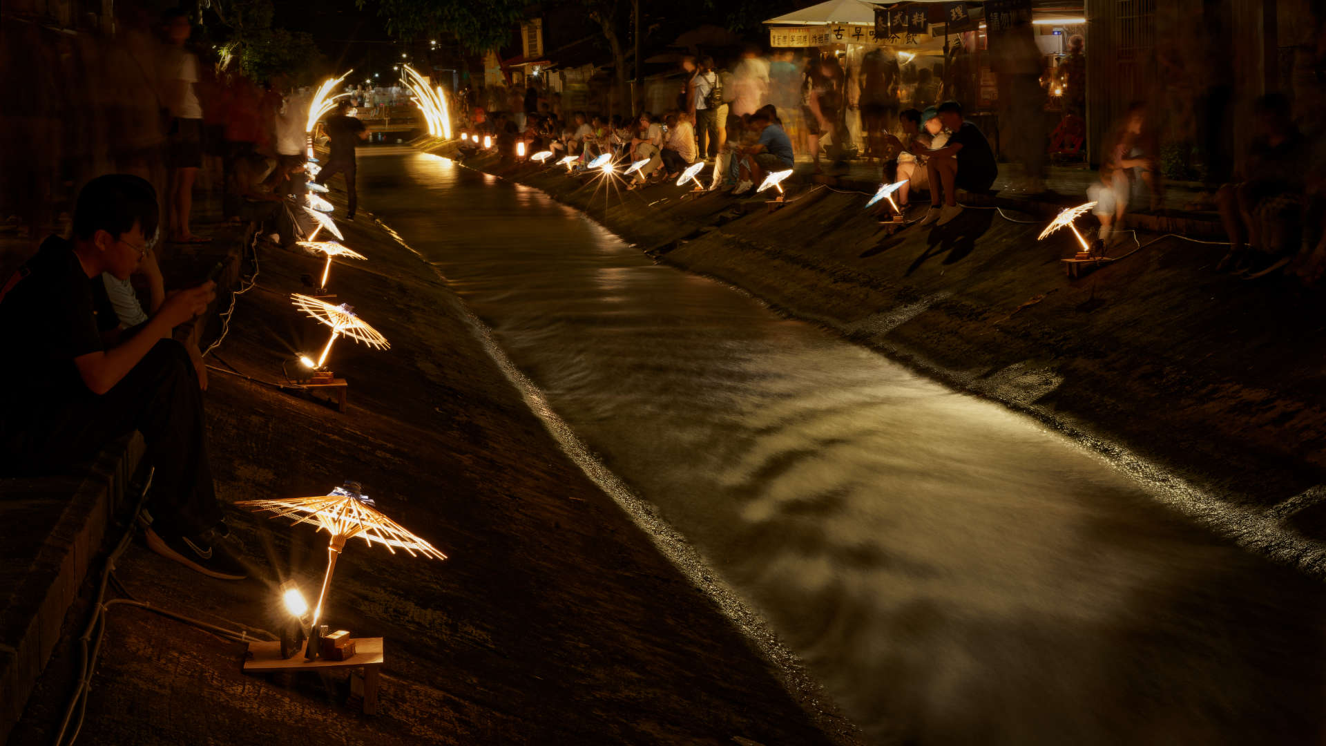 Small wooden umbrellas, illuminated from below, alongside a canal with crowds of people sitting on either side. A few people are walking in the knee-deep water.