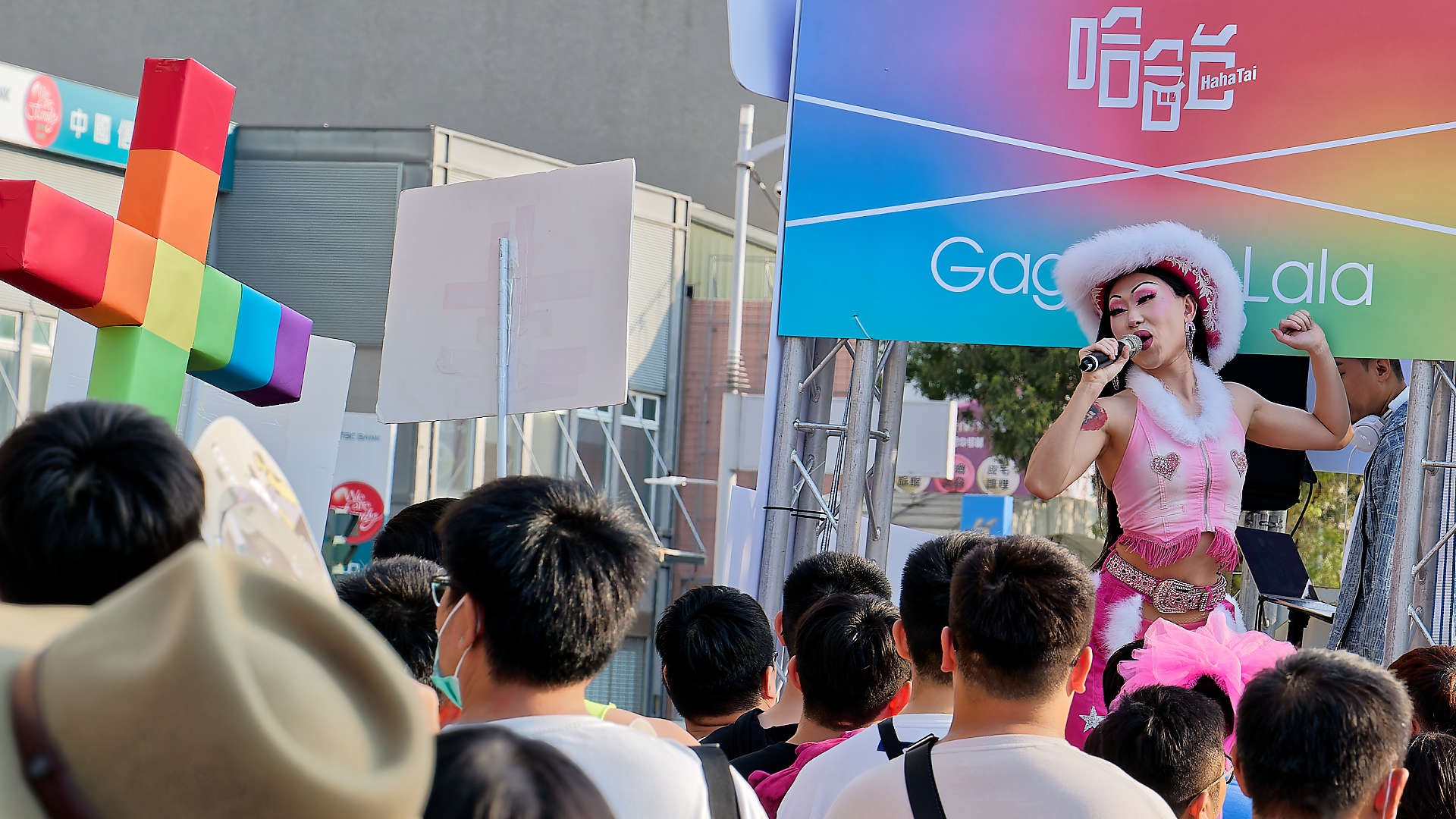 Person singing in front of a crowd. One person in the crowd is holding a rainbow-colored Christian cross.