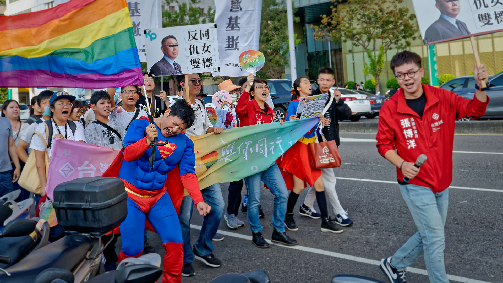 Group of 20 or more people walking together in the Kaohsiung Pride parade. One man is in a Superman costume and holding a large rainbow flag.