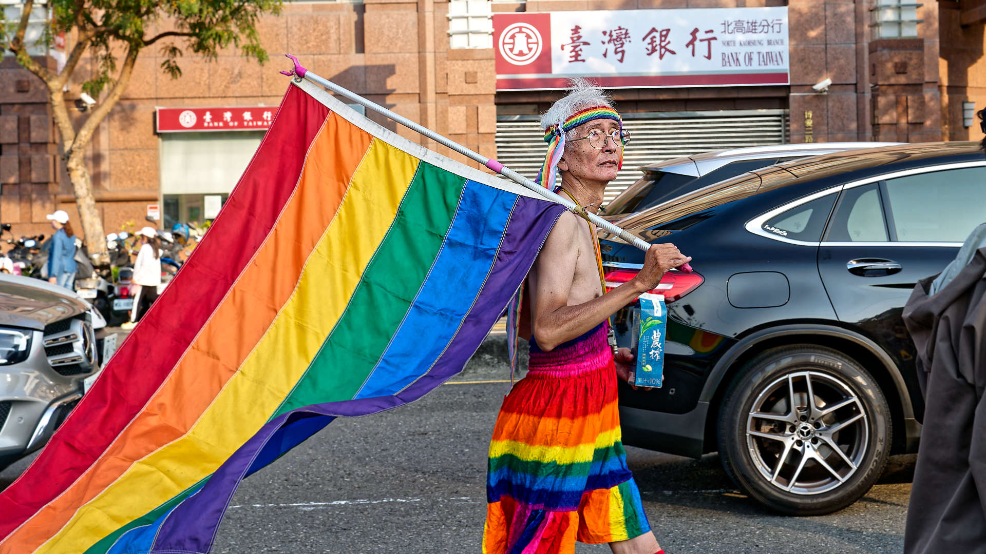 Shirtless elderly man, wearing rainbow-colored pants and carrying a large rainbow flag.