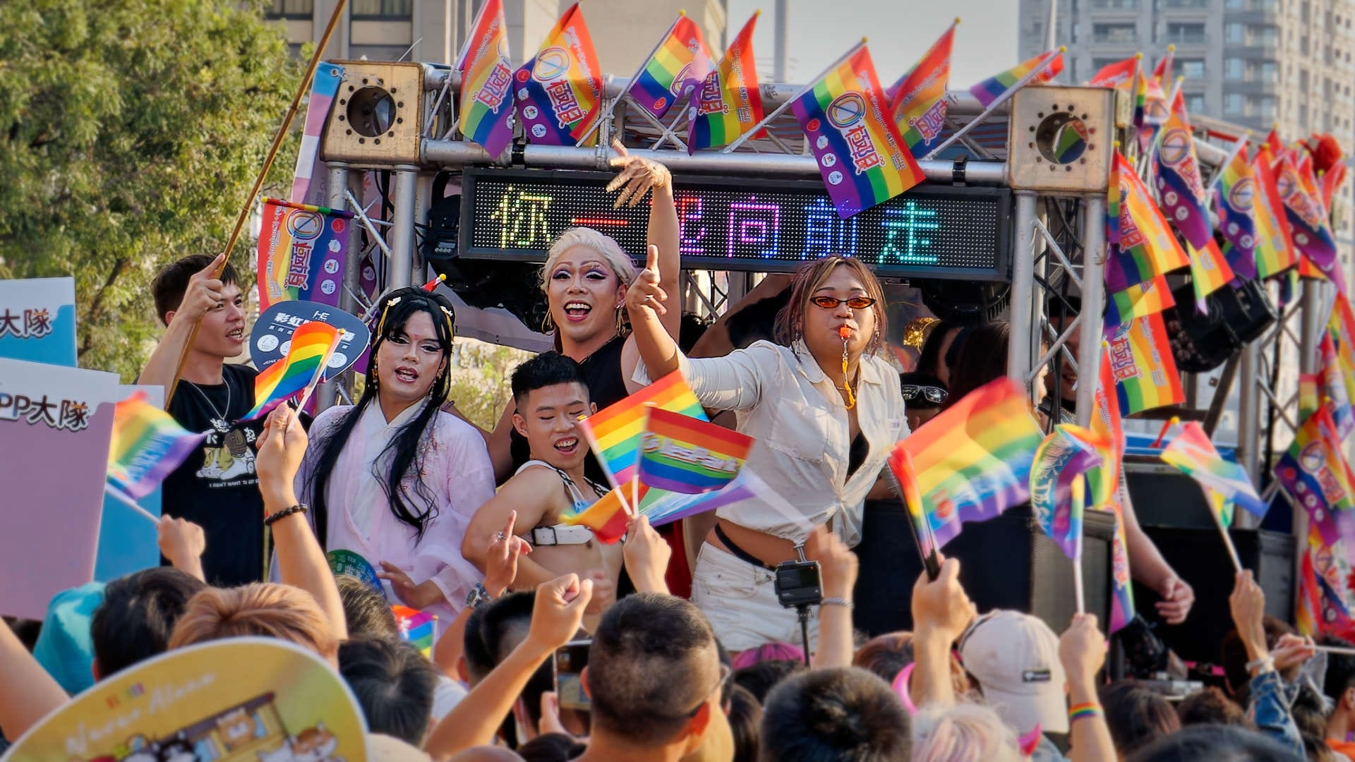 Group of people singing and dancing, surrounded by rainbow flags.