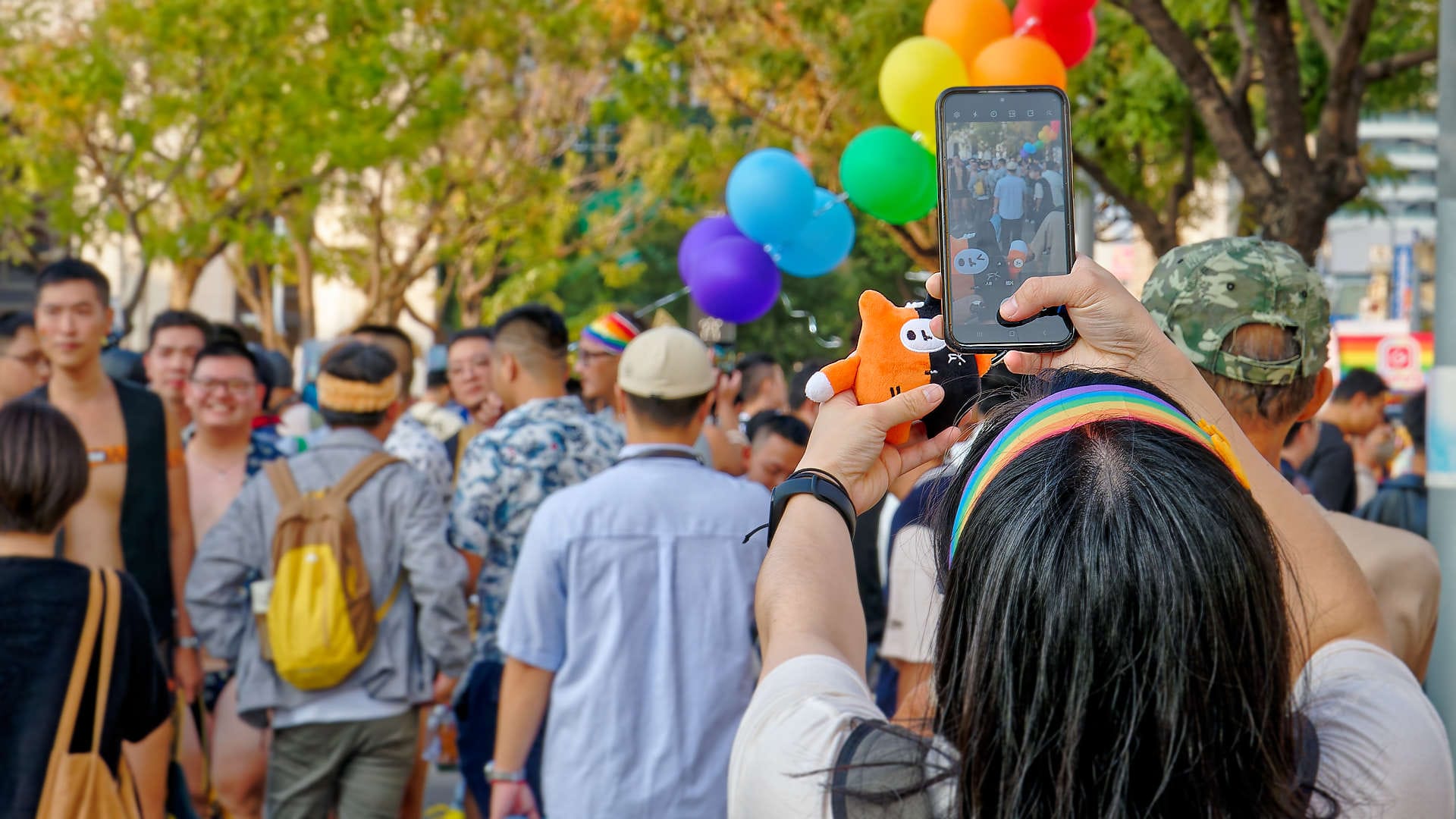 Person taking a photo of a small stuffed toy, with the Kaohsiung Pride crowd in the background.