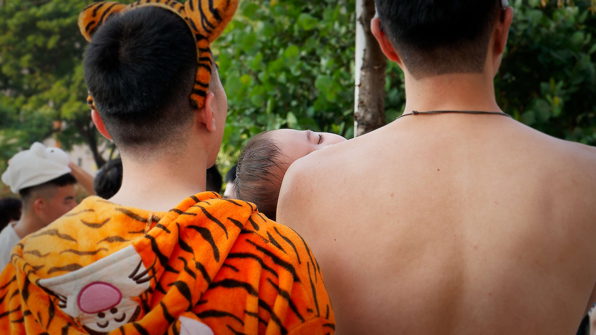 Back view of two men standing close together. The man on the right is holding a sleeping child.