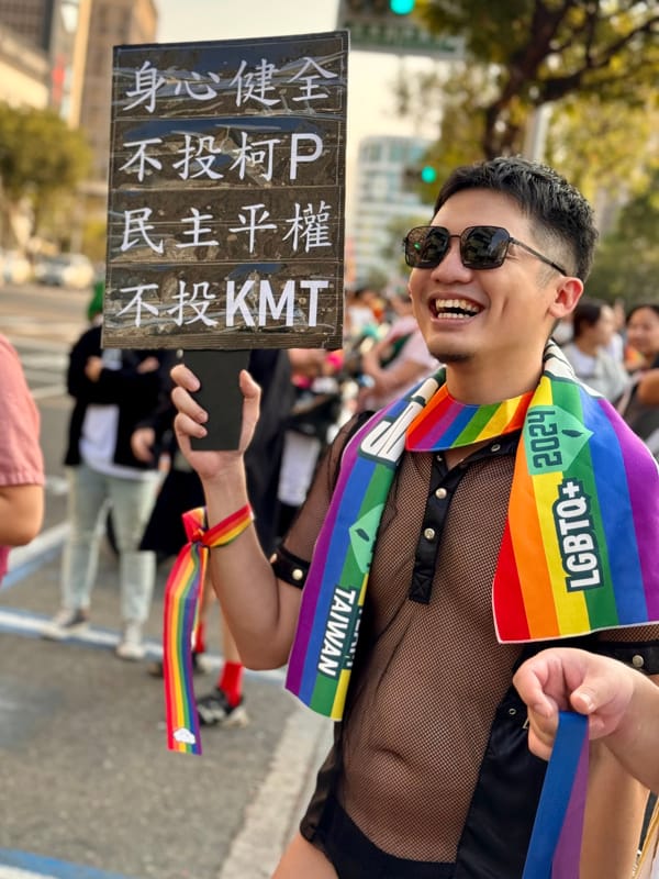 Smiling man draped in a rainbow flag, holding a protest sign.
