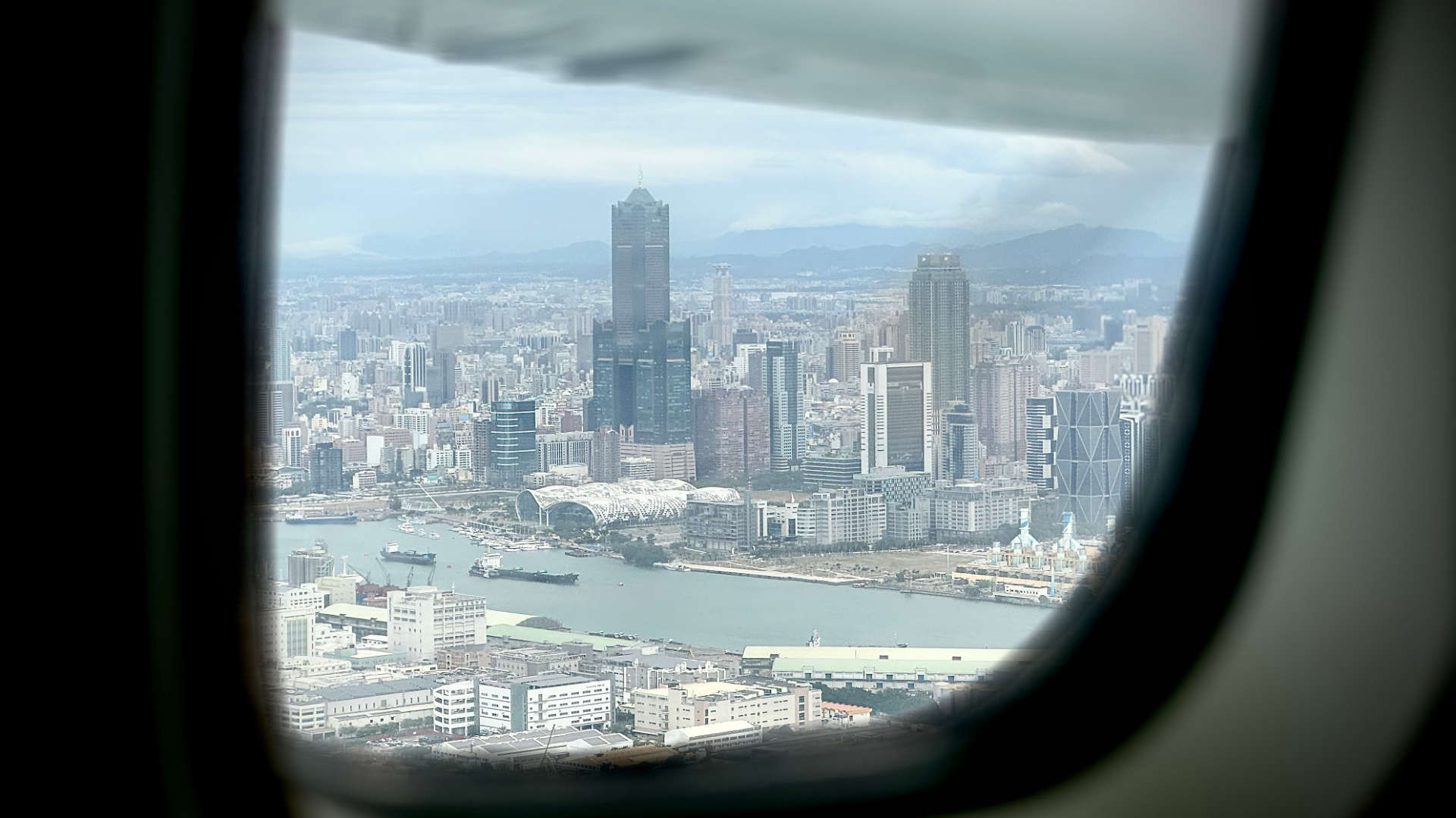 Aerial view of central Kaohsiung, taken through an airplane window. Landmarks including 85 Sky Tower, Kaohsiung Exhibition Center, Kaohsiung Public Library, and China Steel Building, are clearly visible.