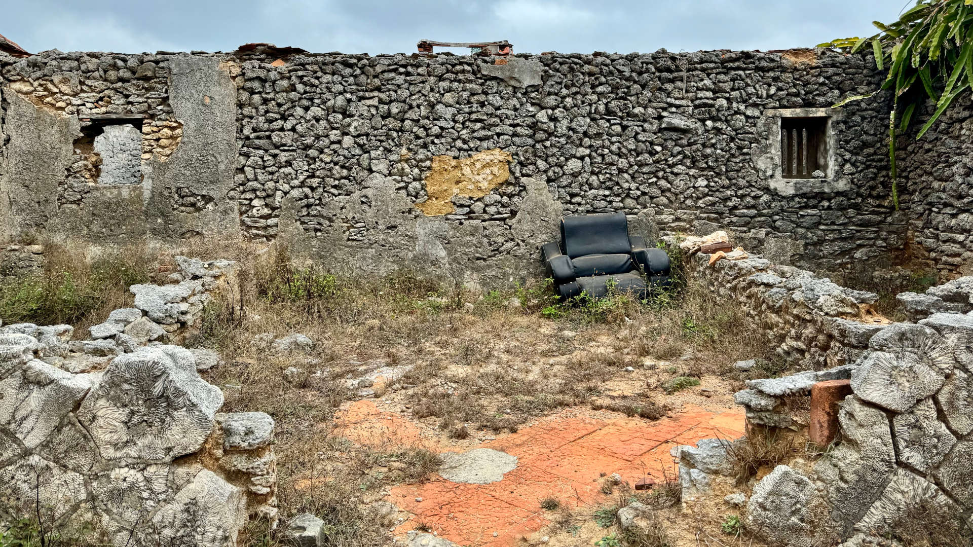 A roofless abandoned building made of coral. A vinyl armchair sits in one corner.