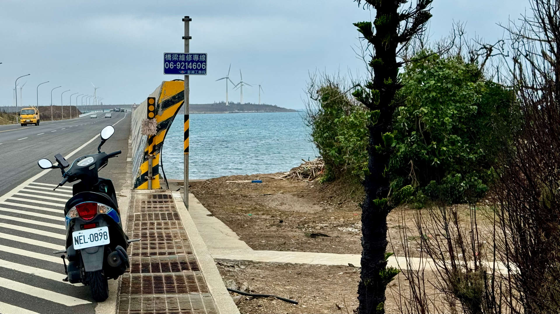 A scooter parked next to a bridge, with wind turbines on the island in the distance.