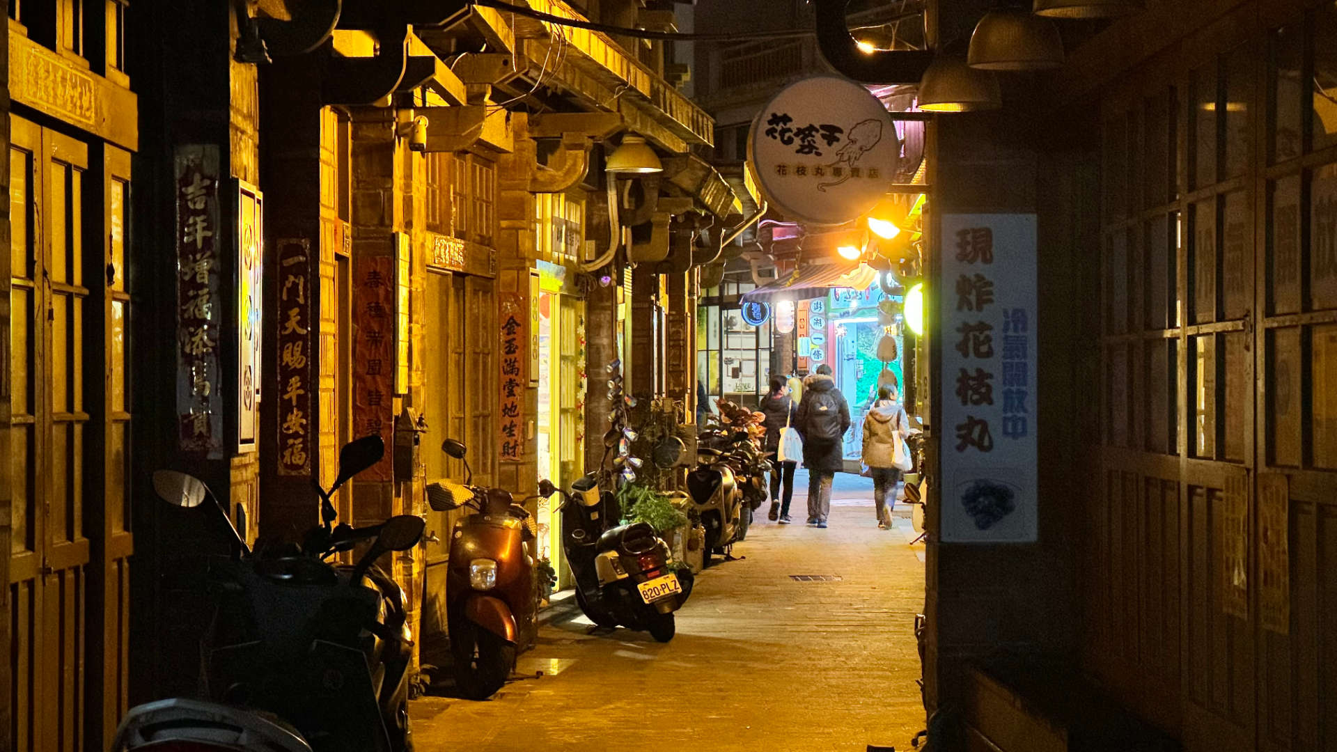 Three people walk along Zhongyang Old Street at night. There are traditional wooden buildings on either side of the narrow street.