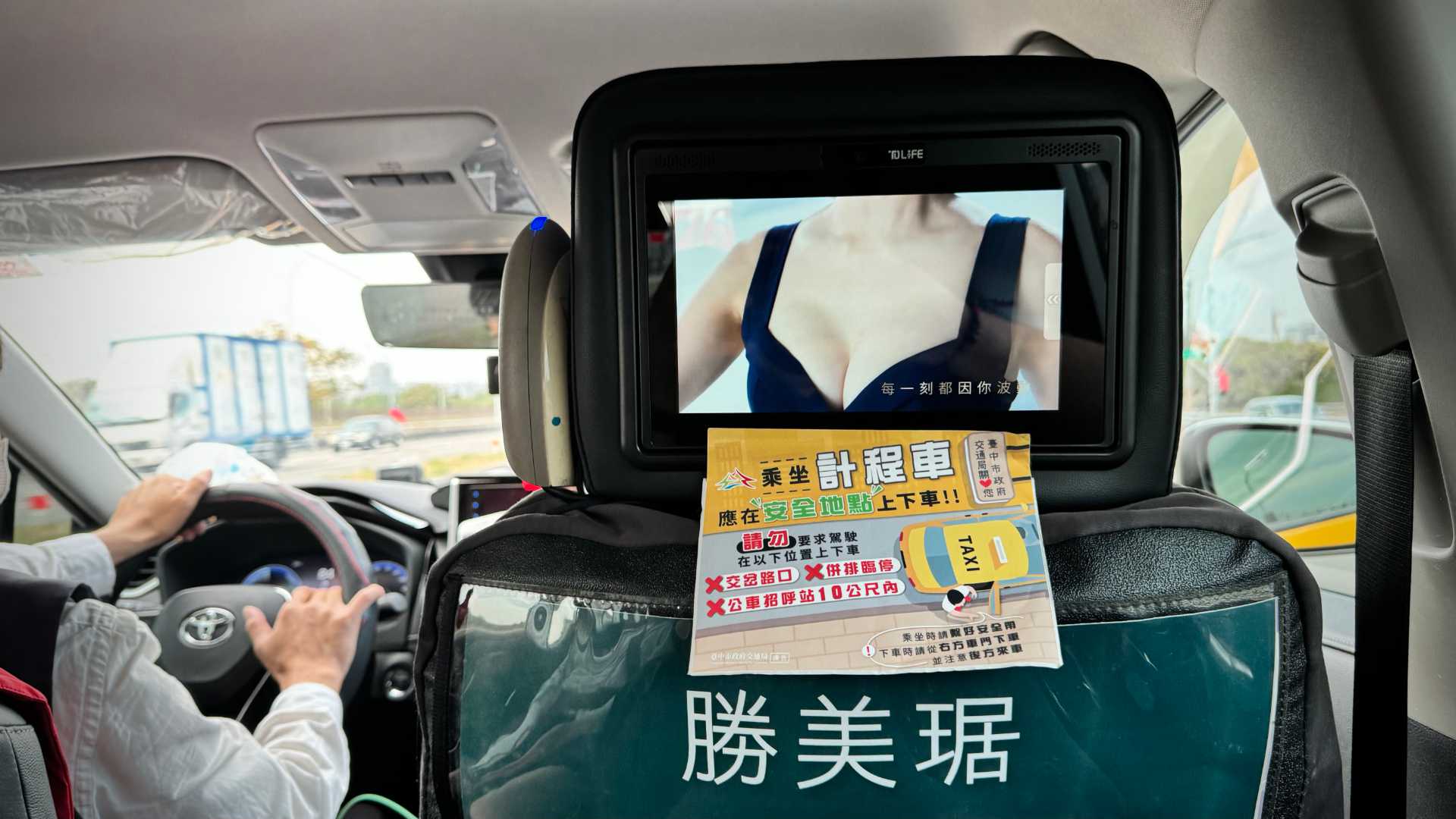 A headrest-mounted TV inside a taxi. The TV screen shows a close-up of a clothed woman’s chest.