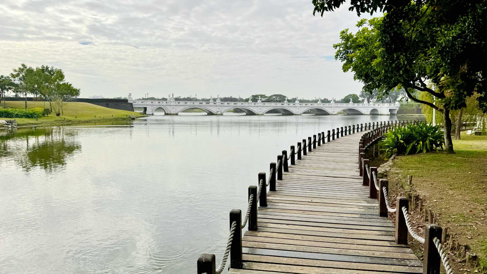 A curved boardwalk next to a lake, with an arched footbridge in the distance.