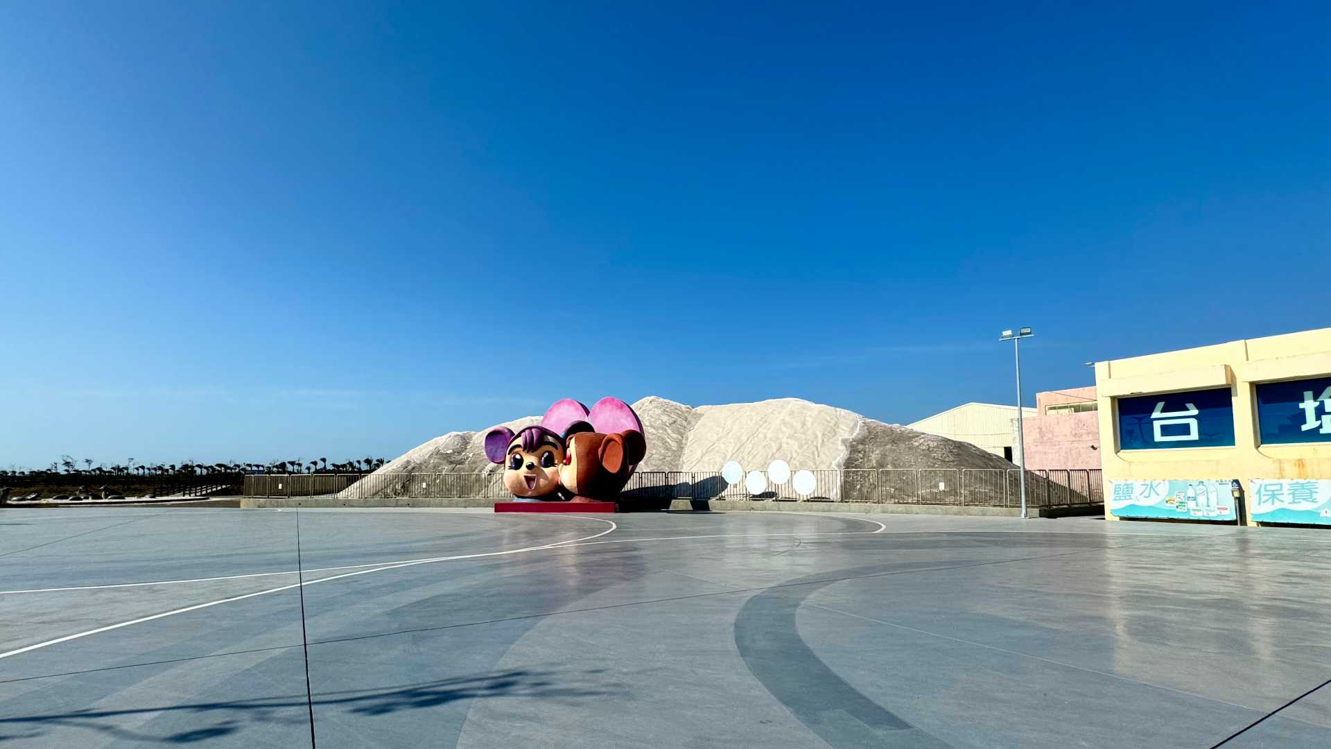 A large pile of salt on the far side of a carpark. The sale pile is maybe 2 stories tall and behind a low fence. In front of the fence are two human-size cartoonish sculptures of mouse-like creatures in love.