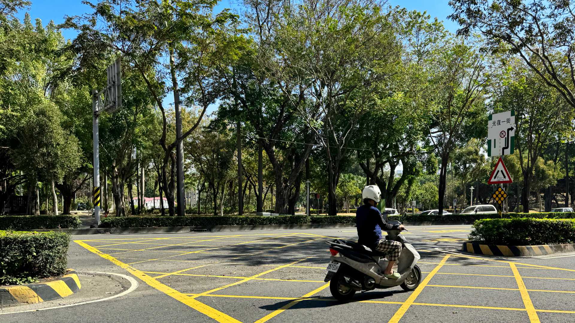 Photo of Central Park taken across a few lanes of traffic. A scooter is driving by in the foreground. The park comprises many tall trees with an arched footbridge in the distance.