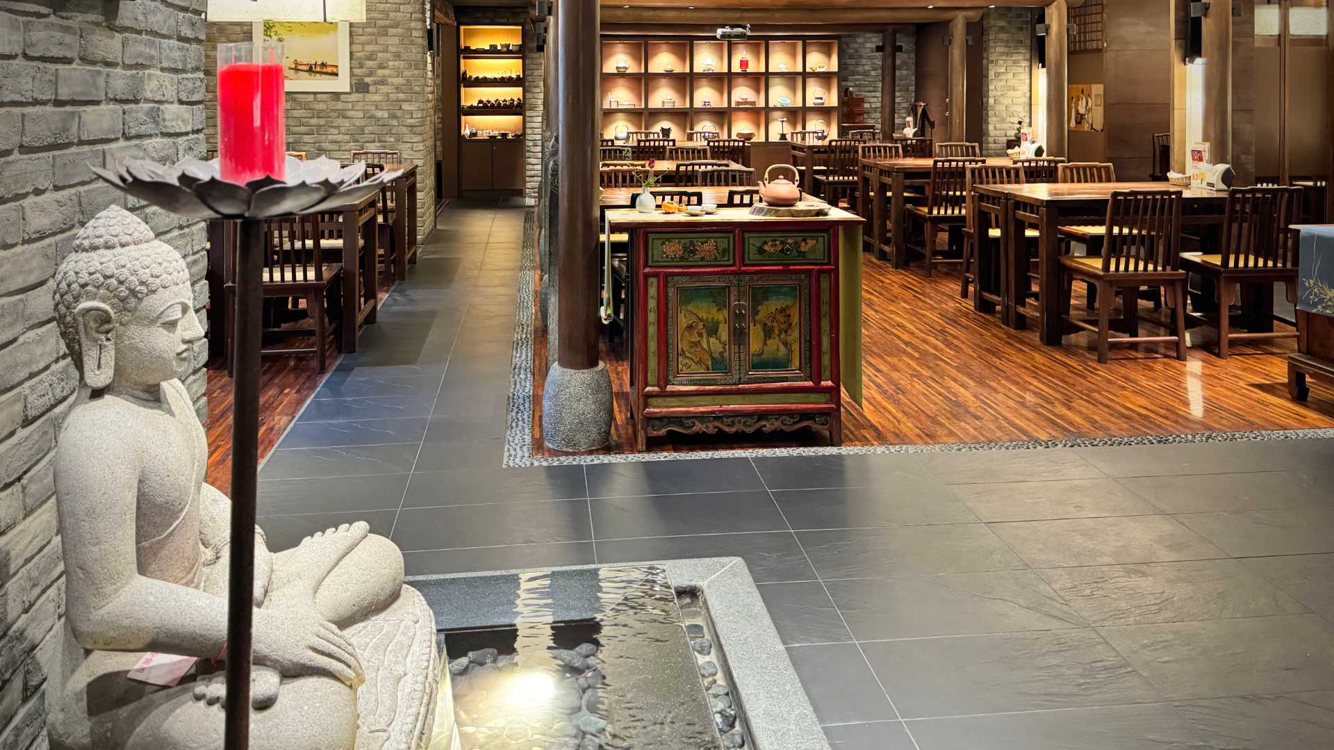 An empty teahouse, with Buddha statue in the foreground. An elaborately-painted side cabinet with a teapot on top is visible in the center of the room, with numerous four-seater wooden tables beyond it.